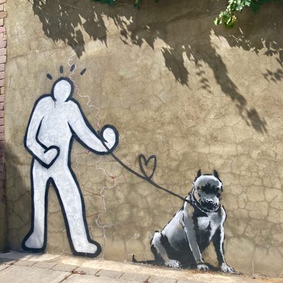 A faceless white figure walking a grey dog, with the leash looping into a heart shape, painted on a beige wall.