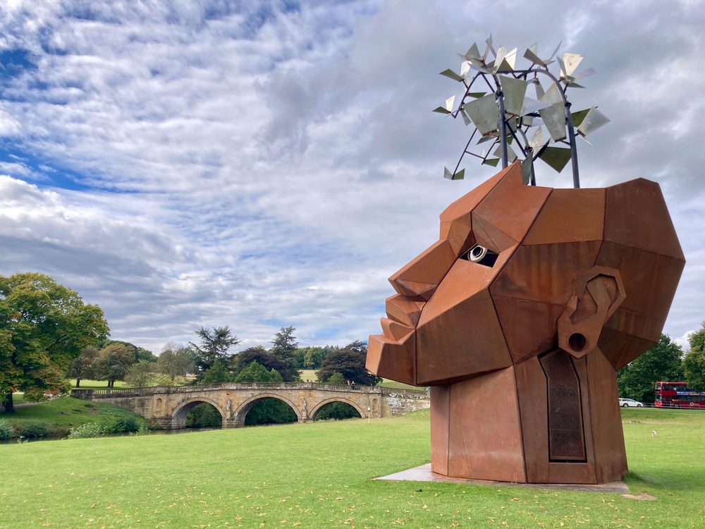A large rust-coloured metal sculpture of a human head sitting on grass, with a stone bridge and parkland behind it.