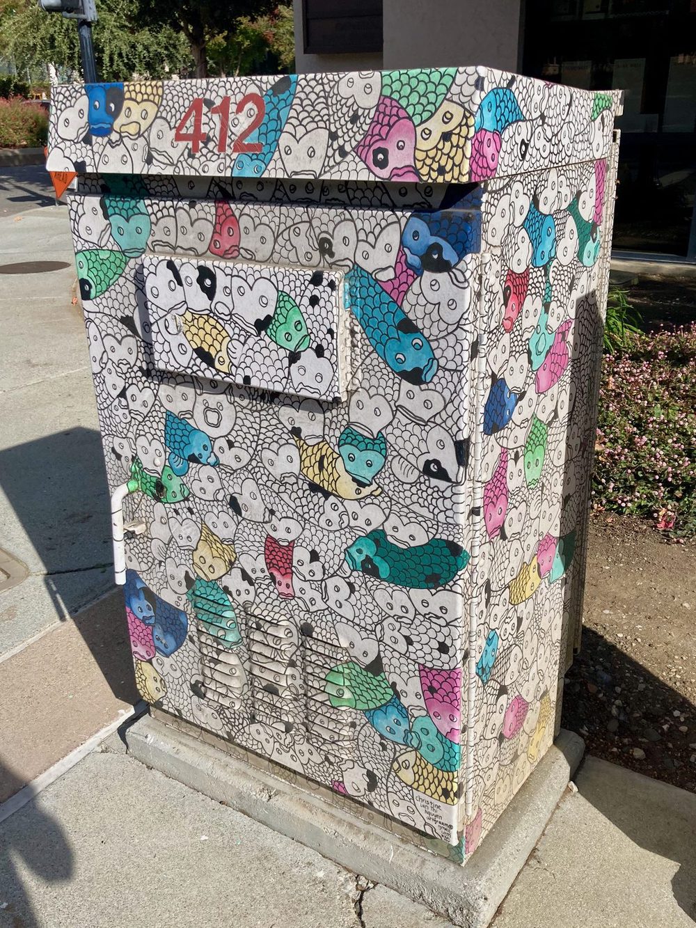 A dense pattern of stylised fish in black outline with blue, pink, green, and yellow accents, painted across a utility box.