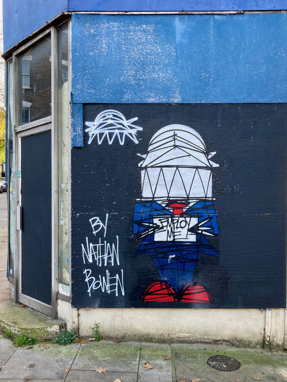A stylised dome-headed figure in blue and red with "Employ me" on its chest, painted on a black wall and signed "By Nathan Bowen".