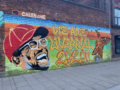 Jürgen Klopp's smiling face in a red cap beside the words "We are Liverpool 2020!!" painted on a brick wall; signed "Caleb.One".