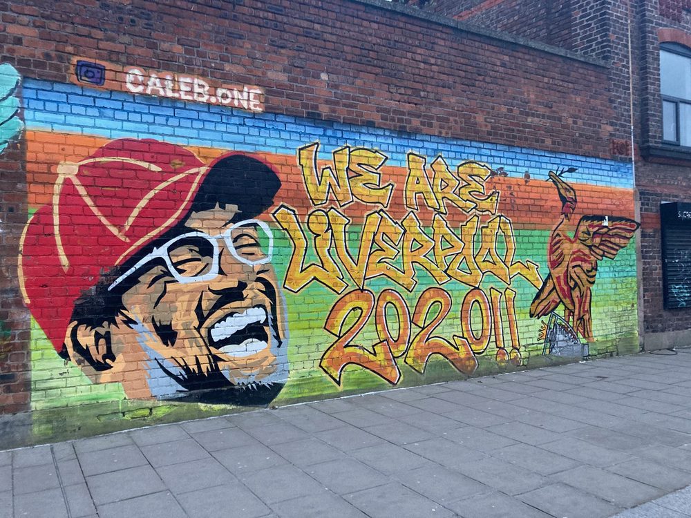 Jürgen Klopp's smiling face in a red cap beside the words "We are Liverpool 2020!!" painted on a brick wall; signed "Caleb.One".