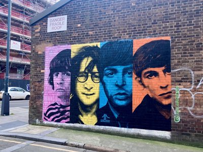 Portraits of the four Beatles — Ringo Starr, John Lennon, Paul McCartney, and George Harrison — in four colourful panels against a black background, painted on a brick wall.