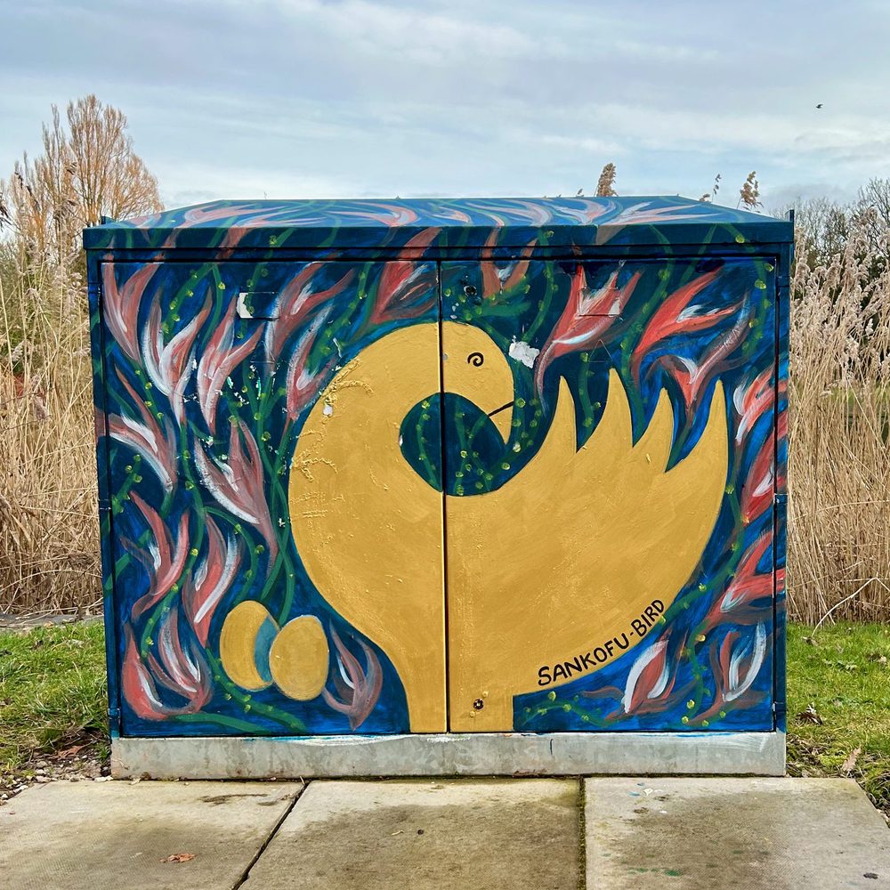 A large golden Sankofa bird among flame-like red and pink patterns, painted on a utility box.
