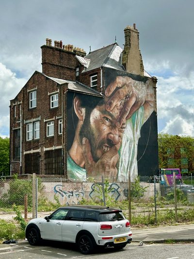 A realistic painted face of a man being pulled in two directions by his own hands, covering the end wall of an old brick building.