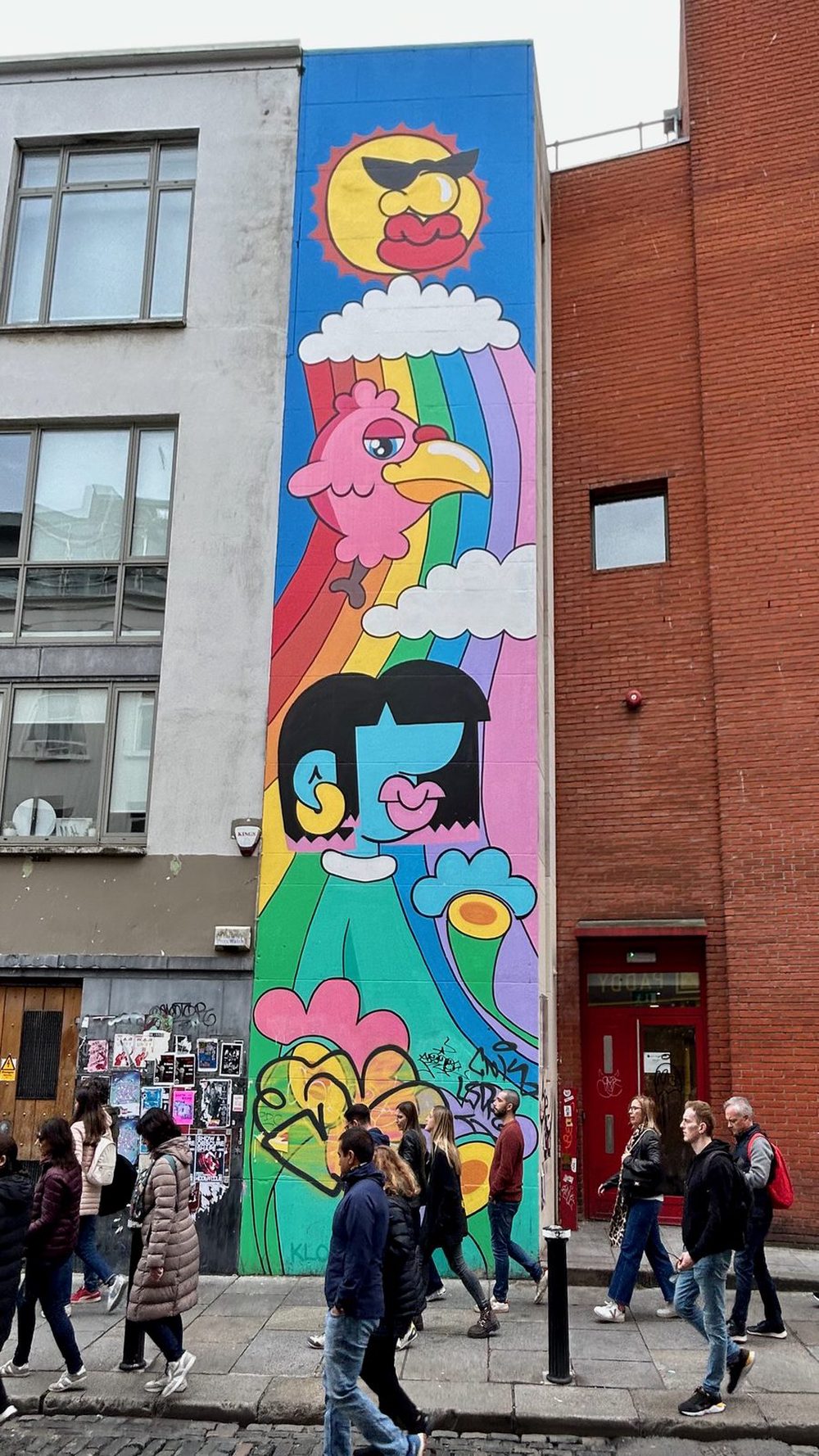 A cartoon sun, a pink cockerel, a rainbow, and a blue-faced figure painted as a tall narrow mural above passing pedestrians.