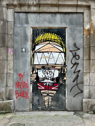 A dome-headed stylised figure with "Paddy Wagon / UFC" on its chest, painted on a metal door surrounded by graffiti; signed "By Nathan Bowen".