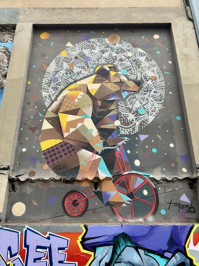 A geometric bear riding a red bicycle, surrounded by dots, triangles, and white circular linework, painted as a geometric mural.
