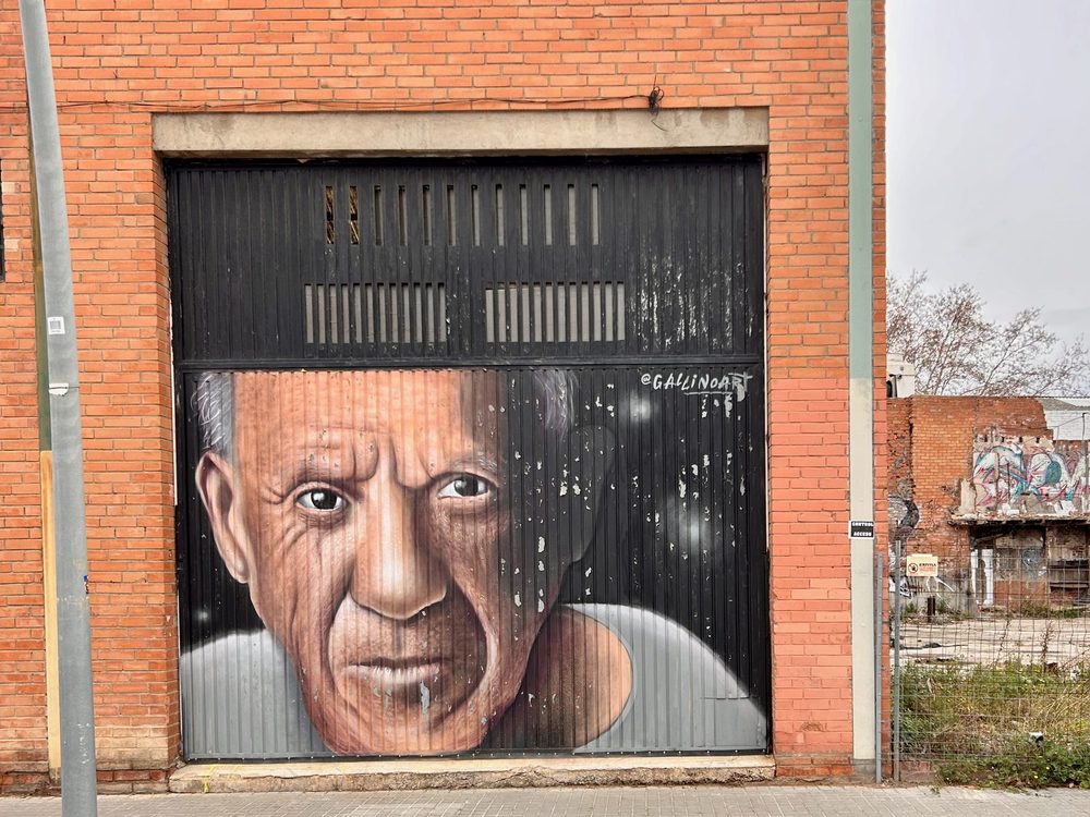 A hyperrealistic portrait of Pablo Picasso painted across a black corrugated door in a red brick building, signed "@GALINDOART".