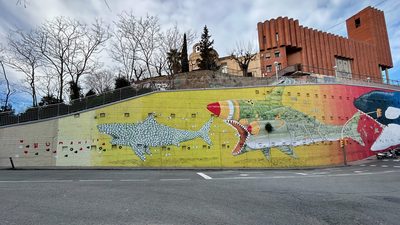 Large sharks — one made of consumer waste, another shaped like a missile — painted across a long yellow roadside wall with the word "Humanity" alongside.