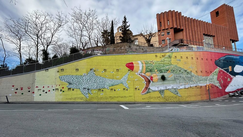 Large sharks — one made of consumer waste, another shaped like a missile — painted across a long yellow roadside wall with the word "Humanity" alongside.