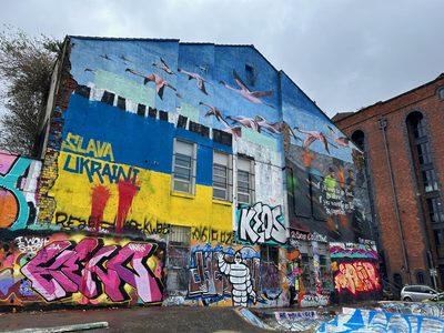 Flying flamingos on a blue upper mural and "Slava Ukraini" painted in yellow and blue below, on a graffiti-covered building wall.
