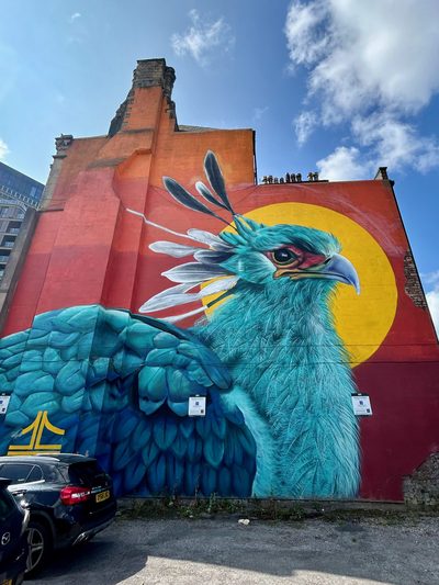 A huge turquoise bird with white head feathers and a yellow sun behind it, painted on a red building wall.