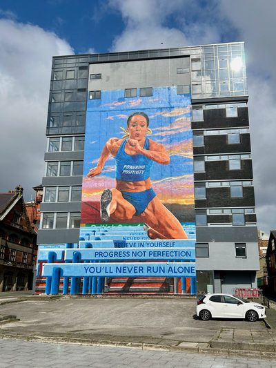 A female hurdler in a blue "Powering Positivity" vest above steps reading "Believe in yourself", "Progress not perfection", and "You'll never run alone.", painted as a tall building mural; signed "Paul Curtis".