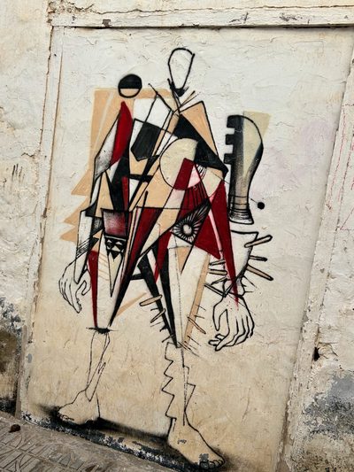 A Cubist-style fragmented humanoid figure in black, red, white, and beige, painted on a rough cream wall.