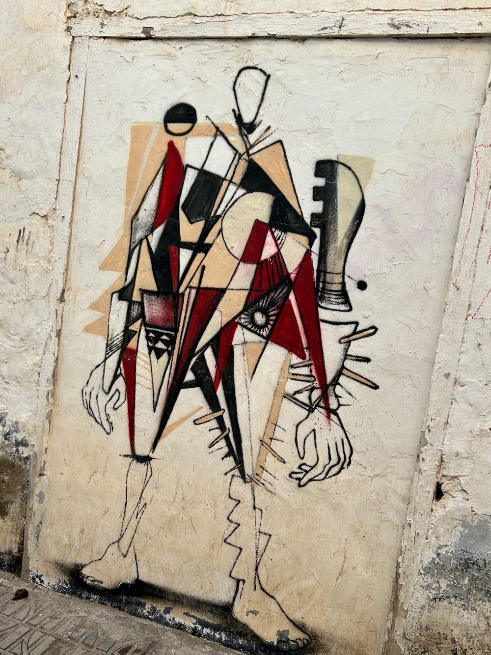 A Cubist-style fragmented humanoid figure in black, red, white, and beige, painted on a rough cream wall.
