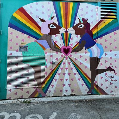 Two unicorn-headed figures touching noses above a pink heart, with rainbow rays behind them, painted on a colourful doorway; signed "Crystal Vielula".