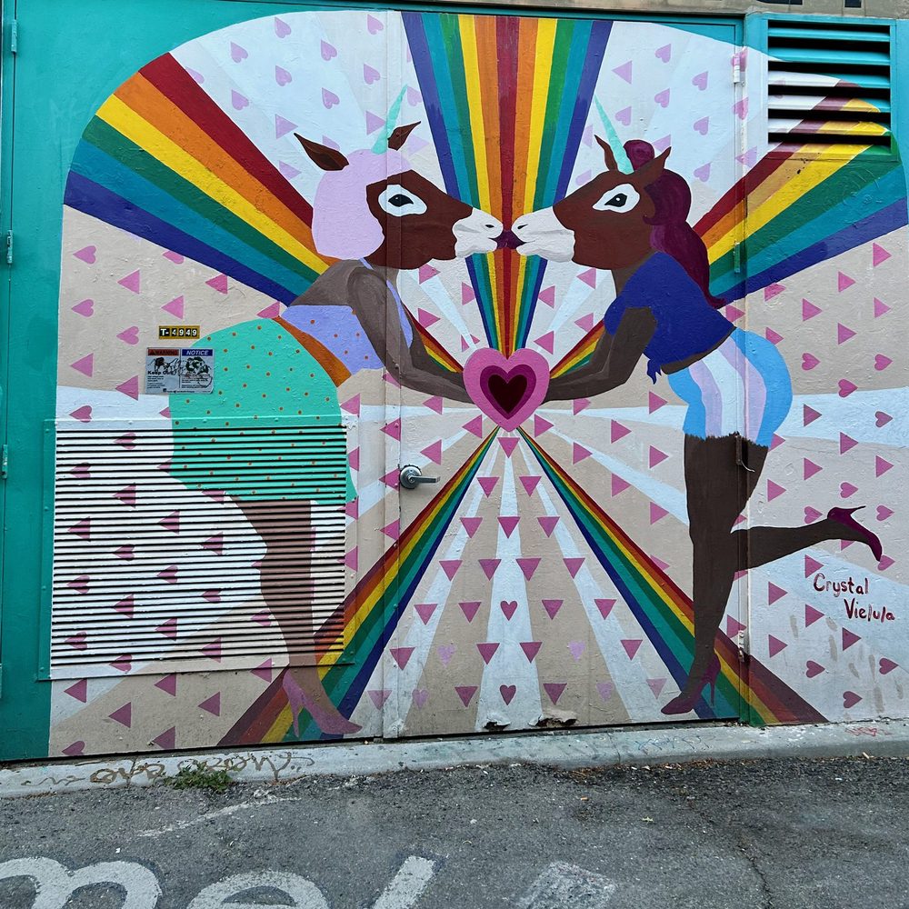 Two unicorn-headed figures touching noses above a pink heart, with rainbow rays behind them, painted on a colourful doorway; signed "Crystal Vielula".