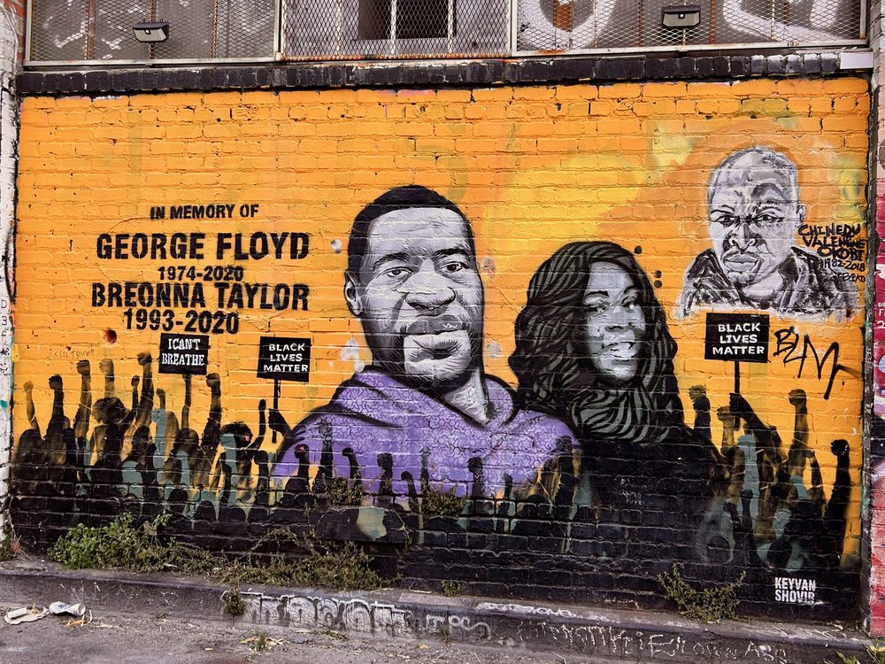 A memorial mural reading "In memory of George Floyd 1974–2020 / Breonna Taylor 1993–2020", with their portraits, raised fists, and "Black Lives Matter" and "I can't breathe" text, on an orange brick wall.