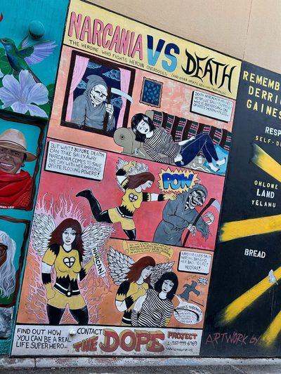 A comic-style mural titled "Narcania vs Death", with a winged superhero in yellow battling a death figure across multiple panels.
