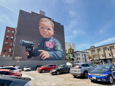 A baby in a police uniform holding a pistol, with a pink toy camera attached to the uniform, painted as a large building mural; signed "BIP".