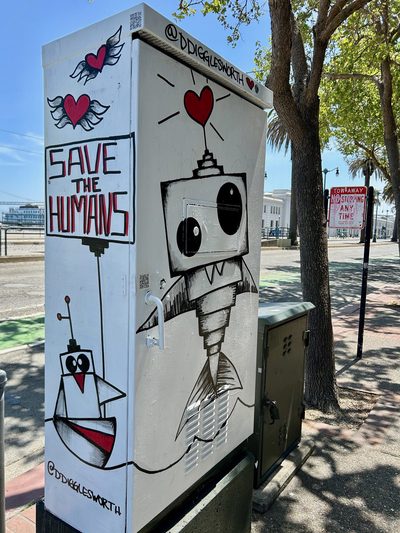 A black-and-white cartoon robot with winged hearts and a sign reading "Save the humans." painted on a white utility box; signed "@ddigglesworth".