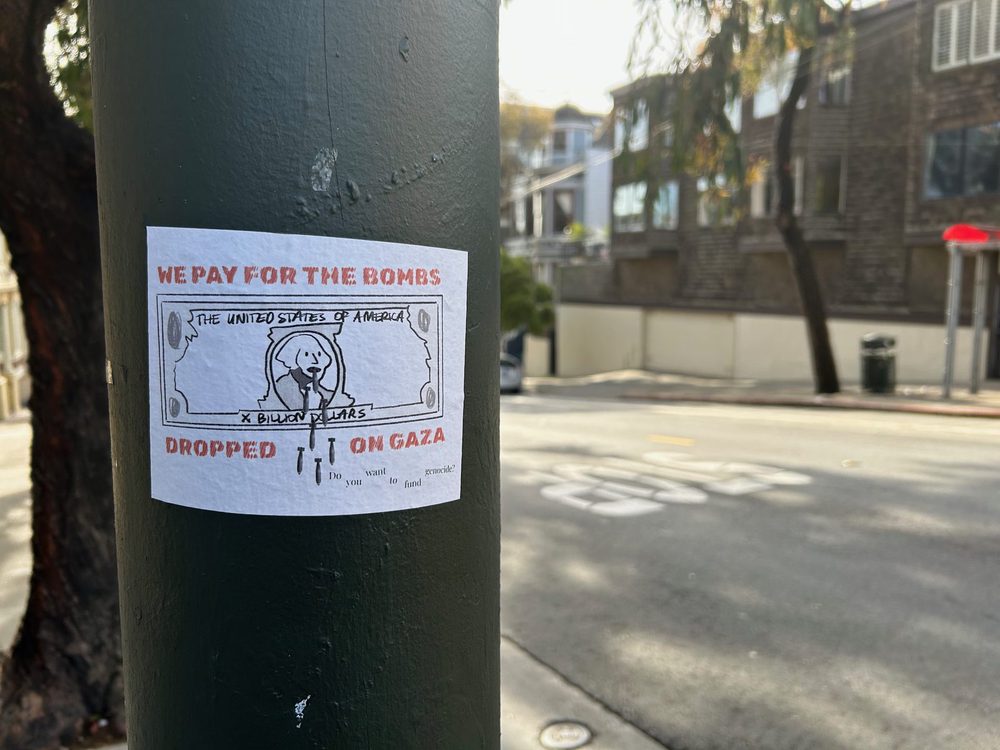 A sticker on a pole reading "We pay for the bombs dropped on Gaza" above a stylised dollar bill marked "X Billion Dollars" and the question "Do you want to fund genocide?"