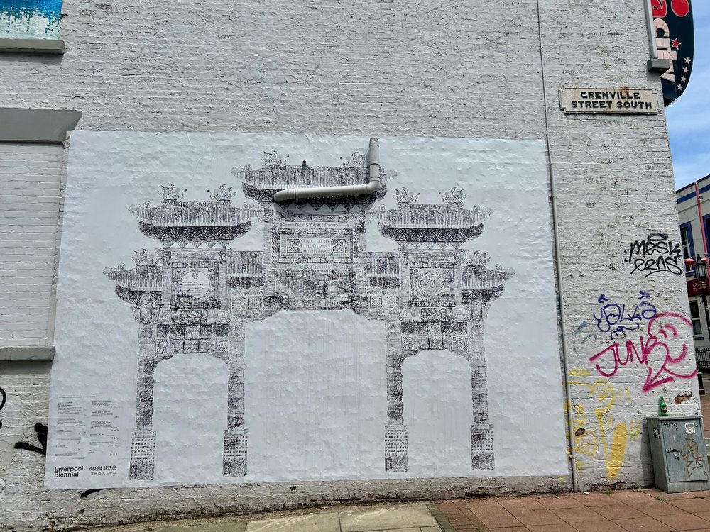 A fine black line drawing of an ornate Chinese paifang gate on a white brick wall, partly crossed by a drainpipe; a Liverpool Biennial and Pagoda Arts commission.