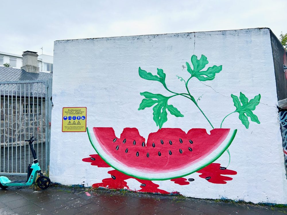 A large watermelon slice with green vines growing from it and red puddles beneath, painted on a white wall.