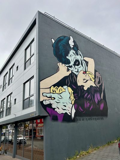 A pale vampire-like figure biting a woman's neck, with Icelandic text reading "Þeim var eg verst er eg unni mest." underneath, painted as a large mural on a grey wall.