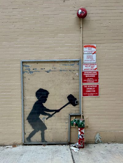 A black silhouette of a child swinging a large mallet, painted on a beige brick wall beside red fire-sprinkler signs.