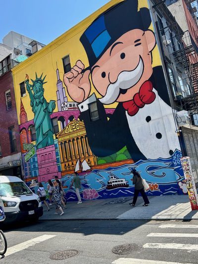 Mr. Monopoly tipping his hat beside New York landmarks including the Statue of Liberty and the Chrysler Building, painted as a bright mural.