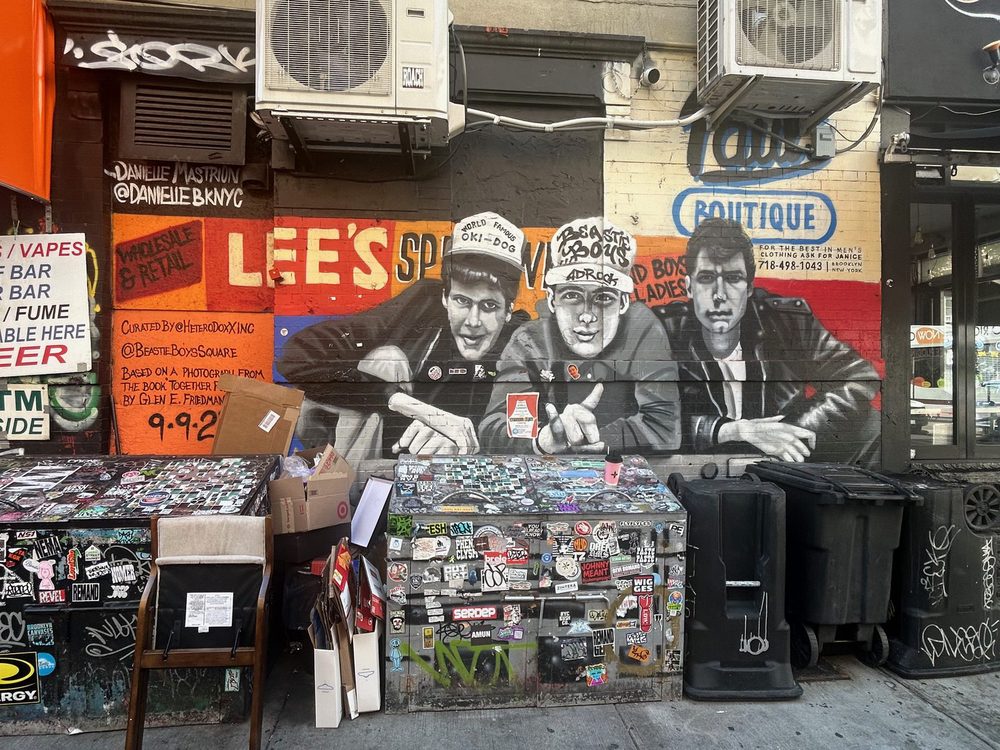 Black-and-white portraits of the Beastie Boys over a red-and-orange "Lee's Sportswear" background with "Paul's Boutique" sign, painted on a storefront.