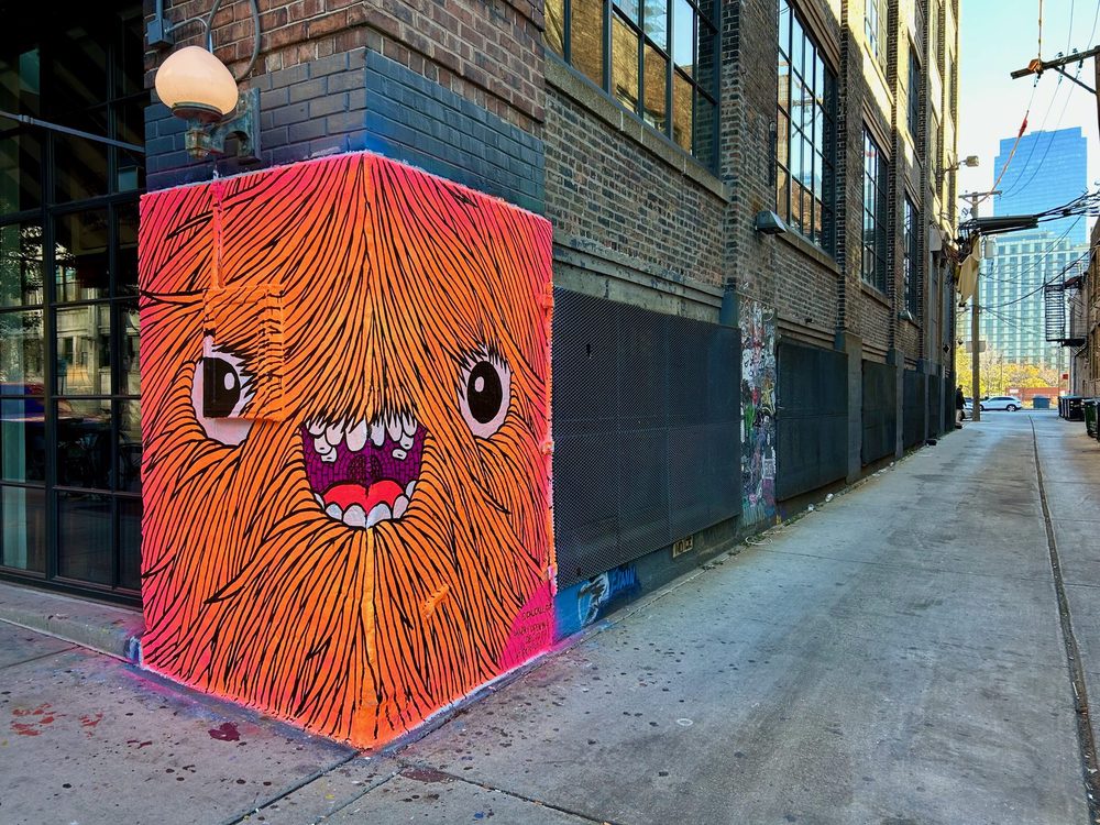 A bright orange shaggy monster face with large eyes and an open toothy mouth, painted across a corner wall.