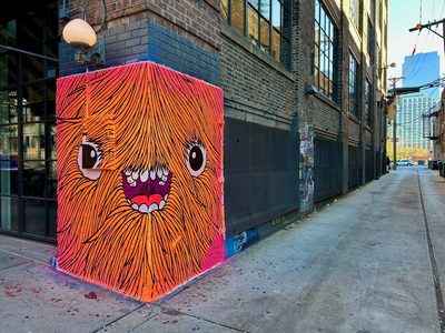 A bright orange shaggy monster face with large eyes and an open toothy mouth, painted across a corner wall.
