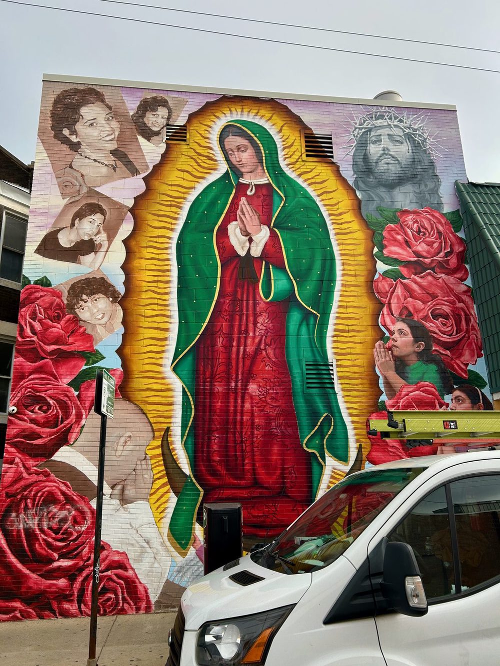 Our Lady of Guadalupe at the centre of a building mural, surrounded by red roses, a crowned Christ figure, and portrait photographs.