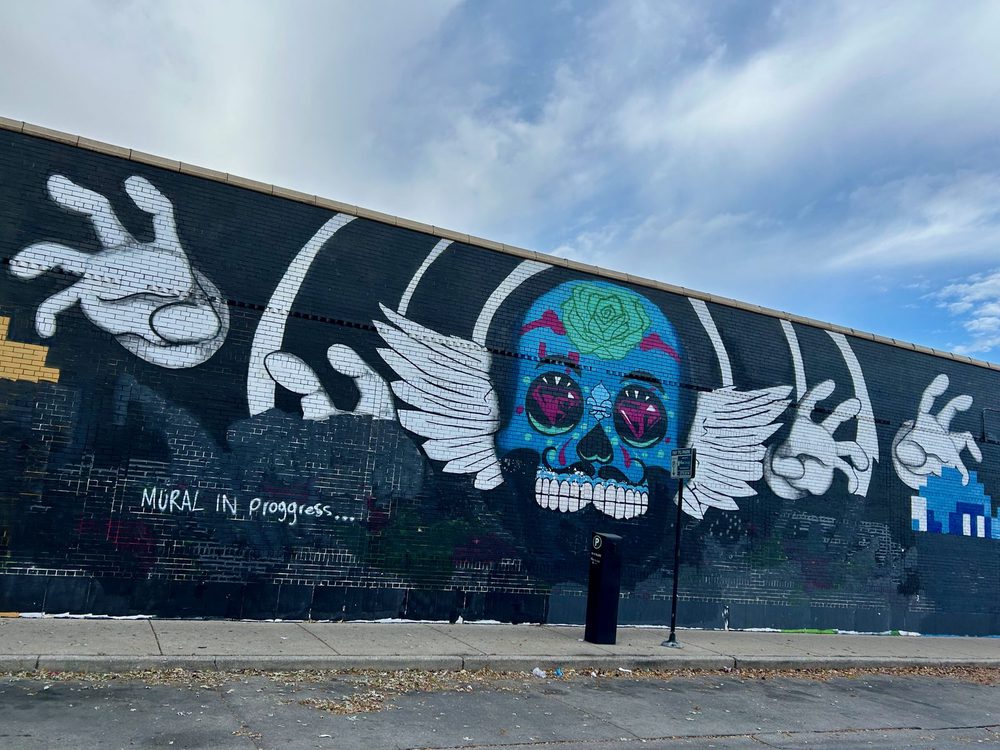 A blue sugar skull, white wings, and oversized white skeleton hands, painted as a mural in progress on a black wall.