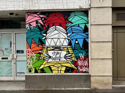 A dome-headed stylised figure with "Stay positive" on its chest, surrounded by colourful geometric shapes, painted on a wall panel; signed "By Nathan Bowen".