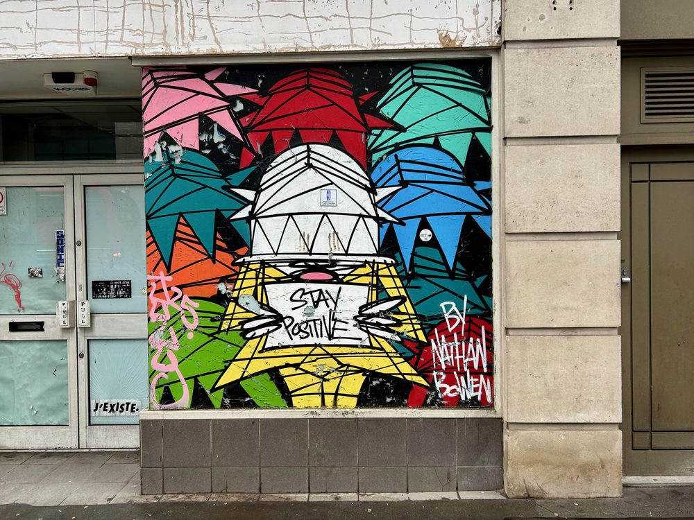 A dome-headed stylised figure with "Stay positive" on its chest, surrounded by colourful geometric shapes, painted on a wall panel; signed "By Nathan Bowen".
