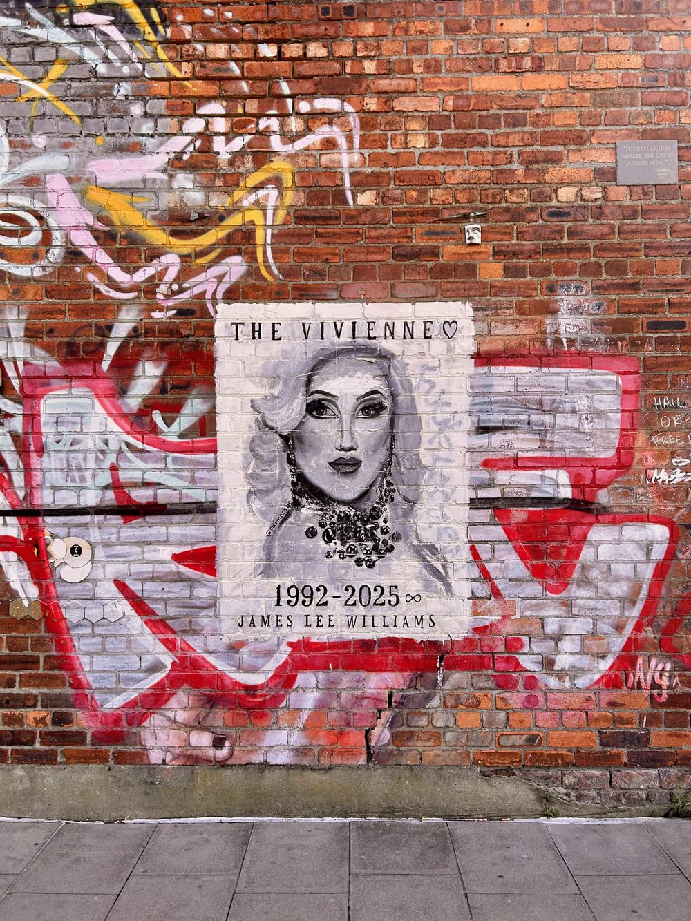A memorial mural on a red-graffitied brick wall: a black-and-white portrait of the drag performer The Vivienne, captioned "The Vivienne" with a heart above, and "1992–2025" and "James Lee Williams" below; signed in the lower right.