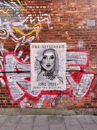 A memorial mural on a red-graffitied brick wall: a black-and-white portrait of the drag performer The Vivienne, captioned "The Vivienne" with a heart above, and "1992–2025" and "James Lee Williams" below; signed in the lower right.