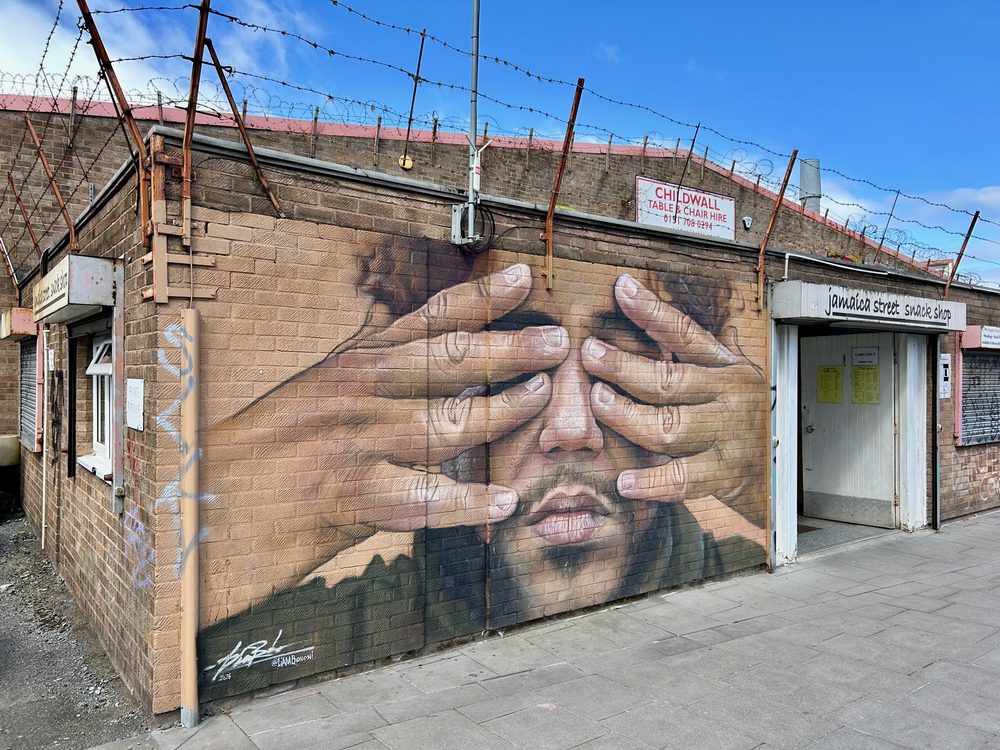 A large photorealistic mural of a man's face, his eyes covered from either side by a pair of hands with fingers splayed across them, painted across the brick wall of a single-storey building topped with barbed wire; an illegible artist's signature in the lower left.