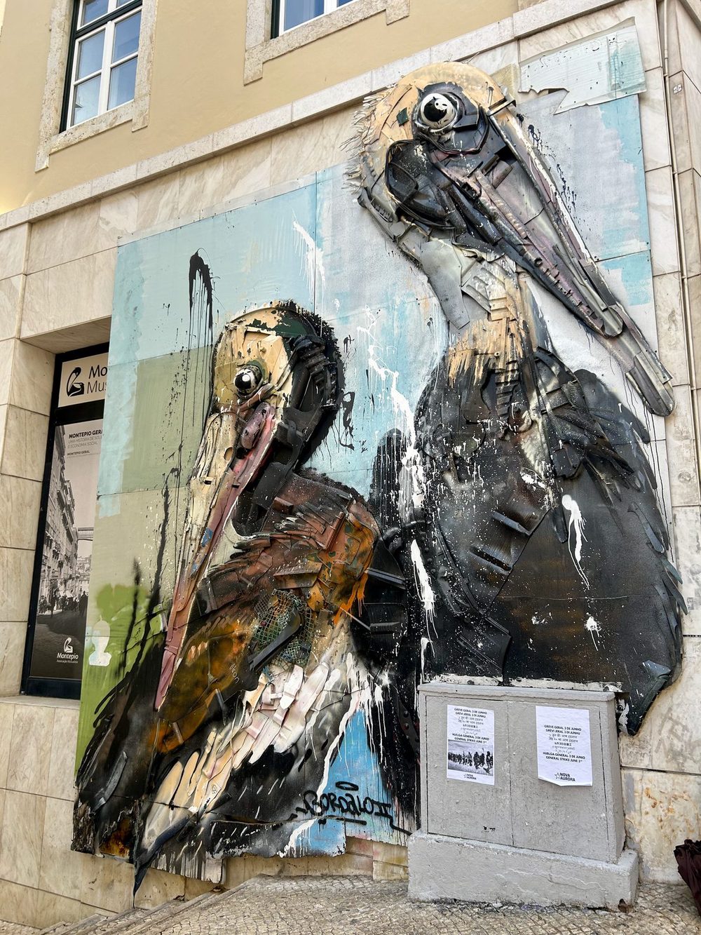 Two large bird sculptures resembling pelicans, built from scrap metal, plastic and other discarded waste, mounted on a stone building wall; signed "Bordalo" in the lower right.