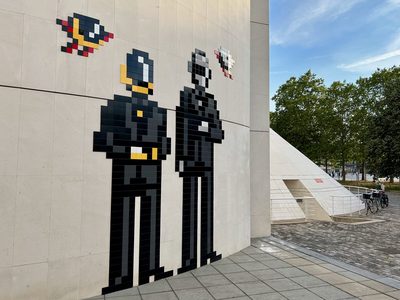A large mosaic of two figures built from black ceramic tiles on a pale concrete wall, dressed in black suits — one with a gold helmet, the other chrome — in the style of Daft Punk. Two smaller pixel-mosaic motifs sit above them. Beyond the wall stand a white pyramidal building, plane trees and parked bicycles.