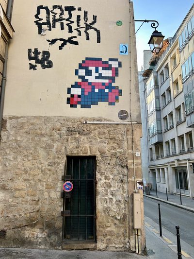 A pixelated mosaic of 8-bit Mario built from ceramic tiles on a beige building corner, high above a stone doorway with a black iron gate and a round no-parking sign. Mario is rendered jumping in red, pink, white, black, and blue tiles. Black stencil graffiti spelling ARTIK is painted above the mosaic alongside small stickers, with a street lamp and Parisian residential facade visible along the narrow street on the right.