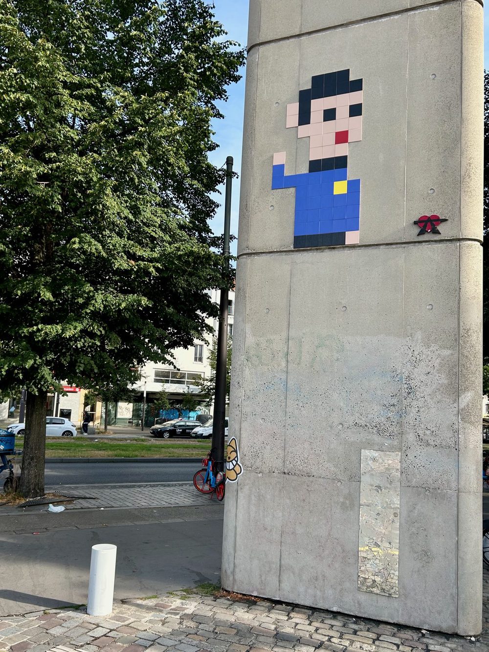A pixelated mosaic of Mr. Spock built from ceramic tiles high on a square concrete pillar, set against a blue shirt with a yellow Starfleet badge. Spock raises his hand in the Vulcan salute. Beside him sits a tiny black-and-red logo sticker, while trees, a street, and parked cars are visible in the background.