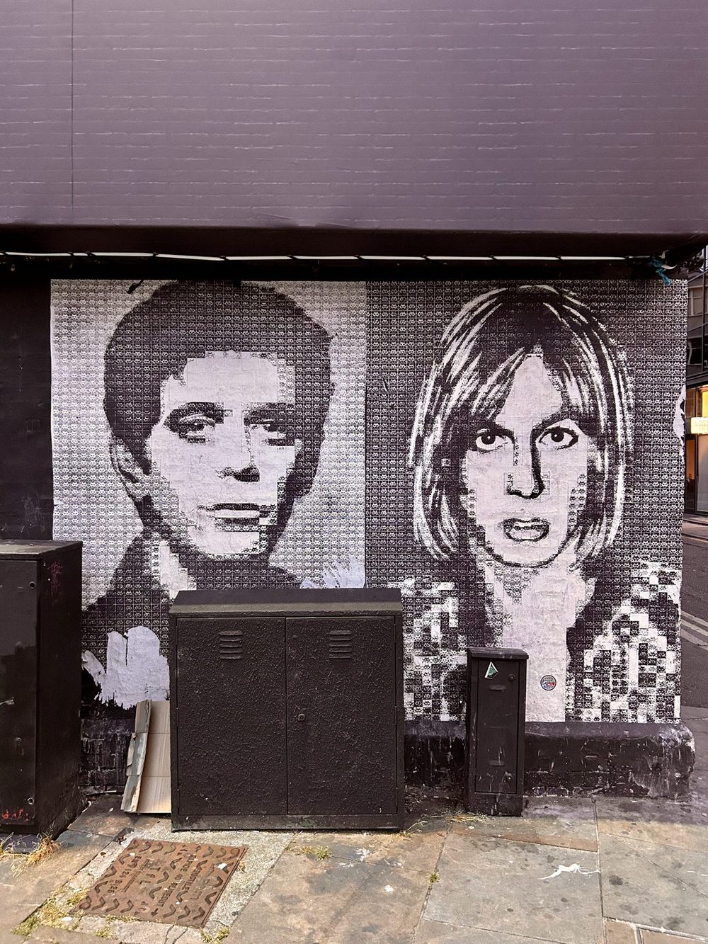 Two large black-and-white paste-up portraits of Lou Reed and Iggy Pop on a tiled wall, partly blocked by utility boxes.