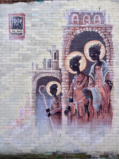 Three figures with dark faces, white pinprick eyes and golden halos, painted on a cream brick wall inside two arched doorways set in painted stone buildings with towers. The tallest wears a pale robe with a rosary and a flower-patterned shawl; the middle figure holds a dark book; a smaller figure sits below holding a crook. Top left, a painted signboard reads 'Hicks' and 'ed_hicks'.