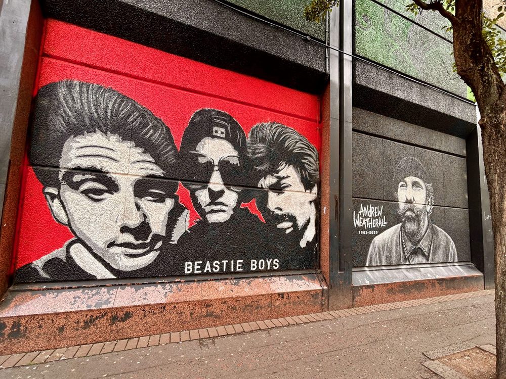 Two painted panels on a dark shopfront. On the left, black-and-white portraits of the three Beastie Boys over a red background, captioned "BEASTIE BOYS". On the right, a greyscale portrait of a bearded man in a flat cap and collared shirt looking upwards, beside the words "Andrew Weatherall" and "1963–2020".